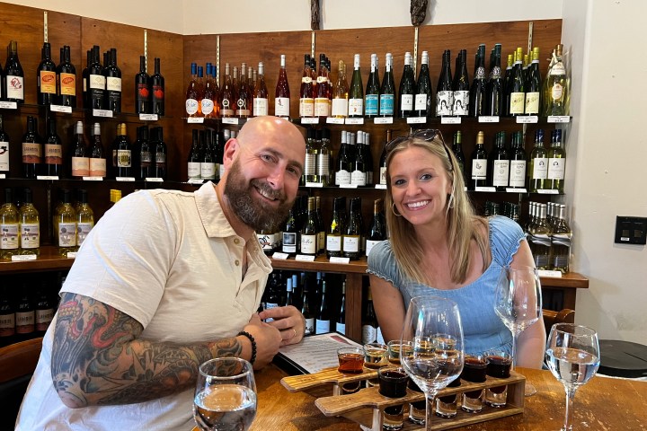 Two people smiling at a table in a wine shop with a wine rack behind them.
