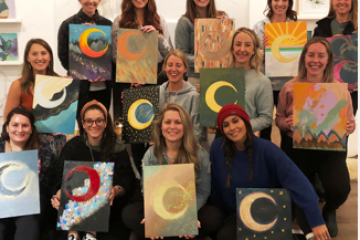 Group of people holding moon-themed paintings, smiling in an art studio setting.
