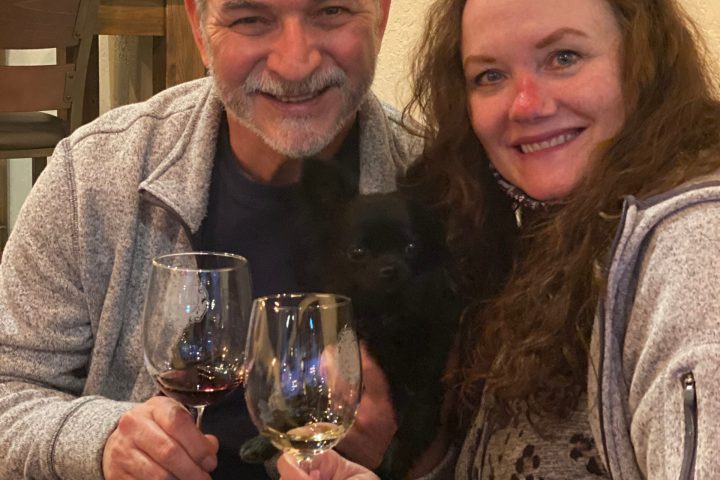 Smiling couple holding wine glasses with a small black dog in their arms indoors.