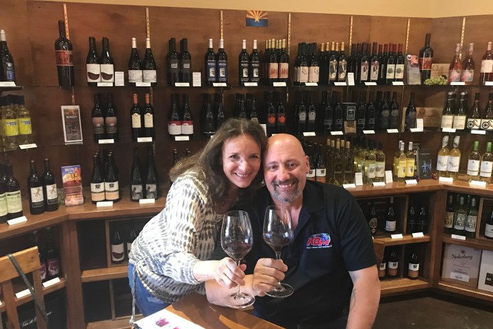 Two people smiling, holding wine glasses in a wine shop with shelves full of bottles.