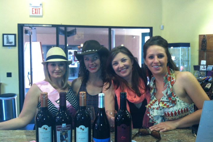 Four women smiling behind a counter with wine bottles in a tasting room.