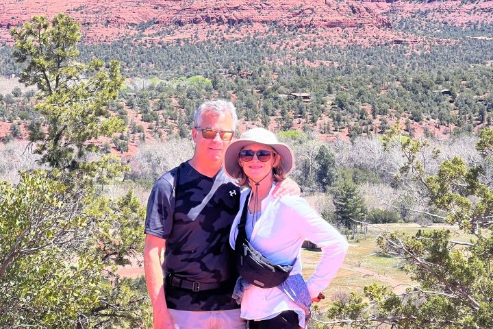 Two people posing in front of red rock formations and trees under a clear blue sky.