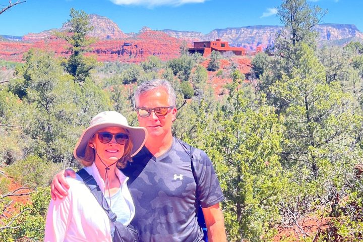 Couple in outdoor clothing stands in a desert landscape with red rocks and green shrubs under a clear blue sky.