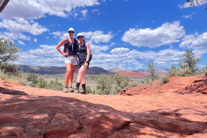 Two people posing under an archway with red rock terrain and blue sky with clouds.