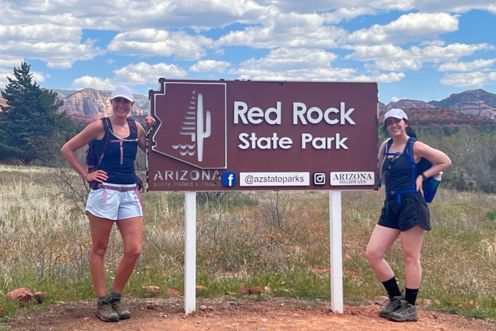 Two people posing by Red Rock State Park sign with backpacks.