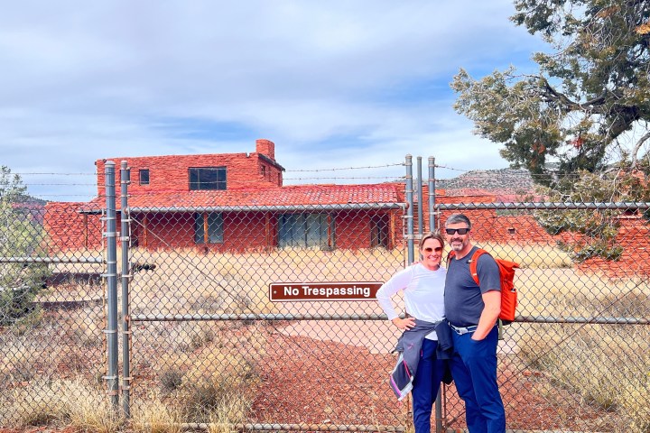 Two people stand near a fence with a 'No Trespassing' sign, in front of a red brick building and trees.