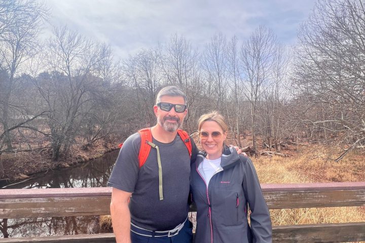 Two people posing on a wooden bridge with bare trees and cloudy sky in the background.