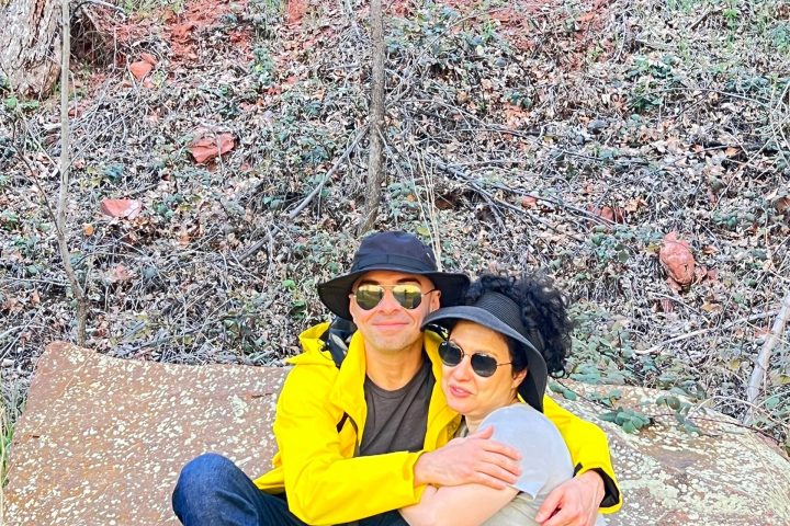 Couple sitting on a rock in a forest, wearing hats and sunglasses, with foliage around.