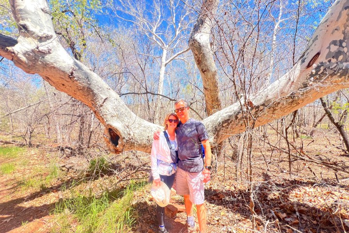 Two people smiling under a large tree branch in a wooded area with blue sky.
