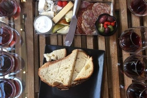Table with wine glasses, sliced bread, cheeses, salami, olives, and dips on plates.