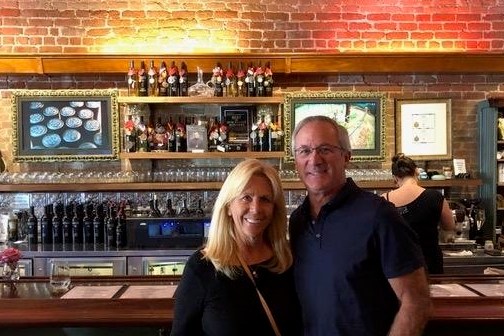 A smiling couple standing in front of a bar with bottles and glasses.