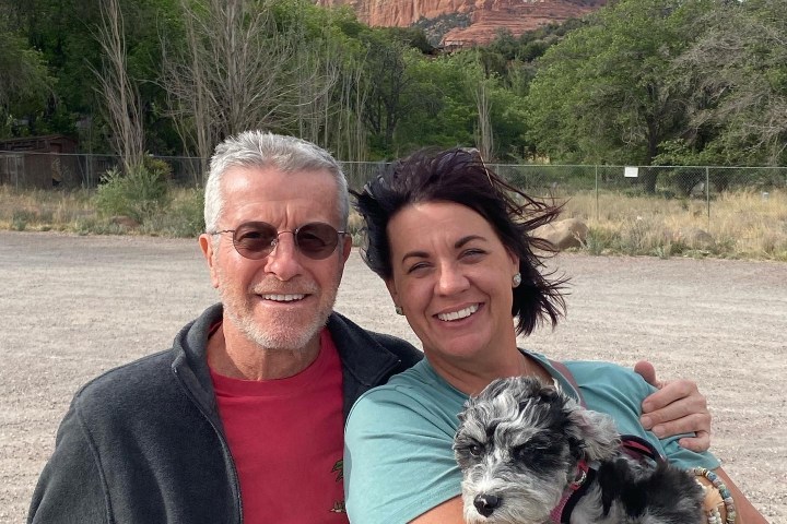 Two people smiling with a dog, set against a backdrop of red rock formations and trees.