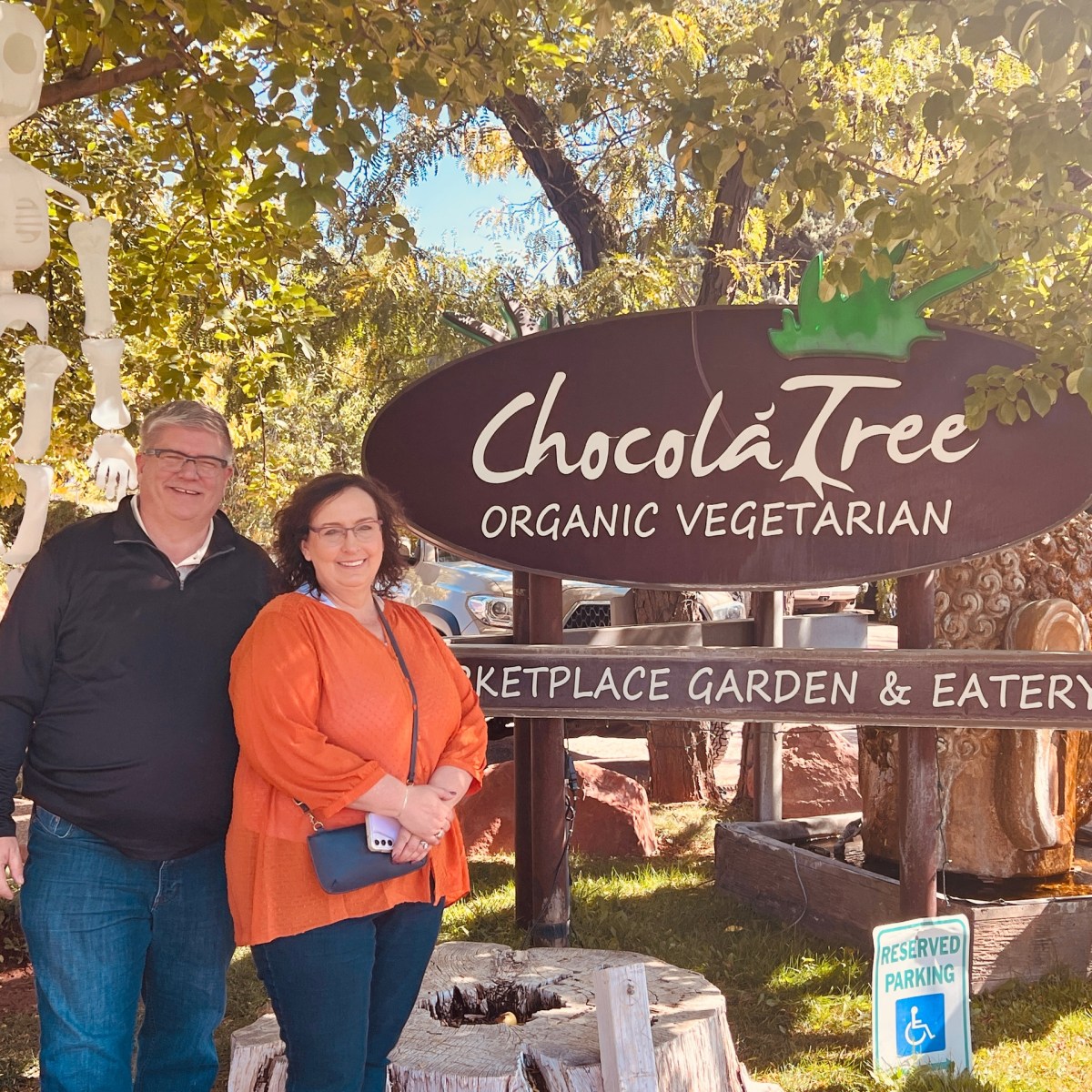 Two people standing beside ChocoláTree Organic Vegetarian sign with a skeleton decoration hanging nearby.
