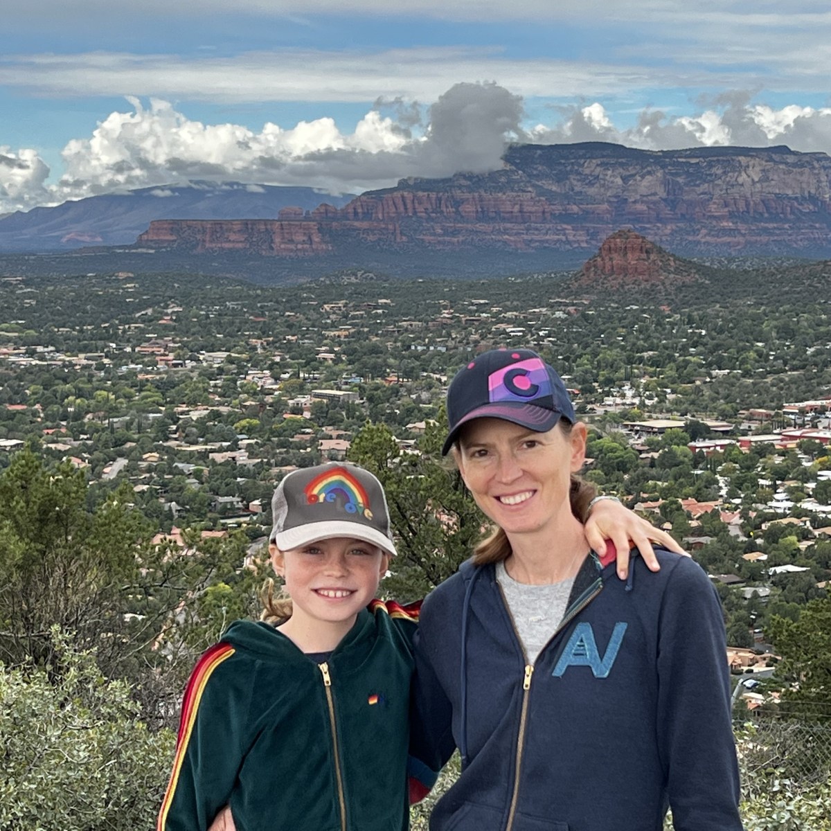 Two people pose with arms around each other, against a backdrop of mountainous landscape and cloudy sky.