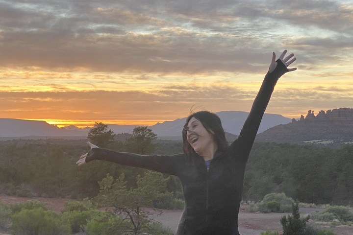 Person joyfully posing with arms up in front of a sunset over a desert landscape.