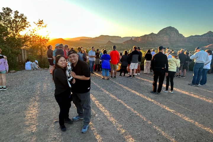 Crowd gathers at sunset to view scenic mountains, with a smiling couple in the foreground.
