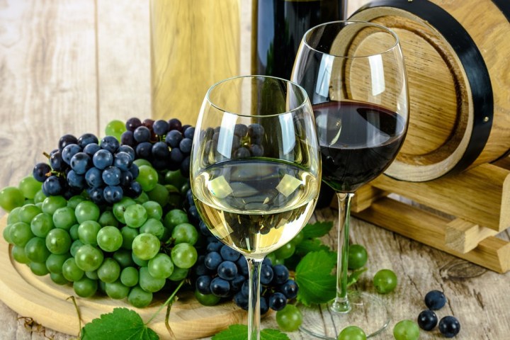 Two wine glasses with white and red wine, grapes, and a barrel on a wooden table.