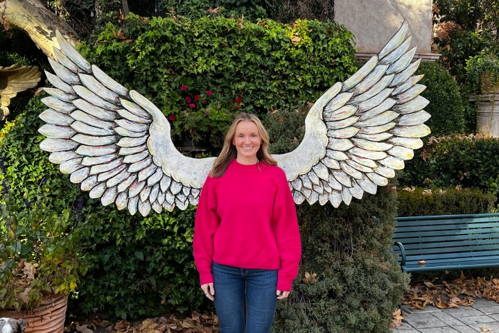 Woman in pink sweater stands in front of decorative wings in a garden.
