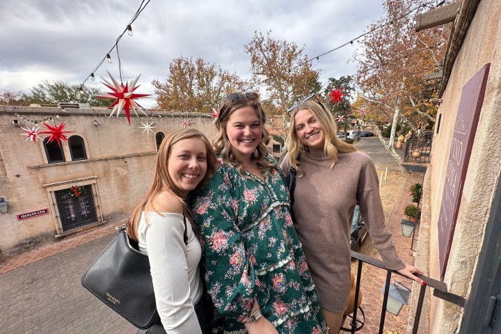 Three women smiling on a balcony with festive decorations and autumn trees in the background.