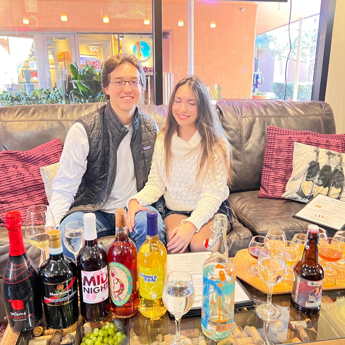 Two people seated on a sofa with assorted beverages on a table in front of them.