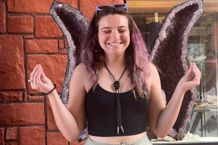 Smiling person posing with amethyst wings against a brick wall backdrop.