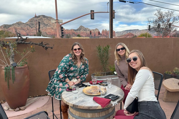 Three women sit at an outdoor table with a mountain view in the background.