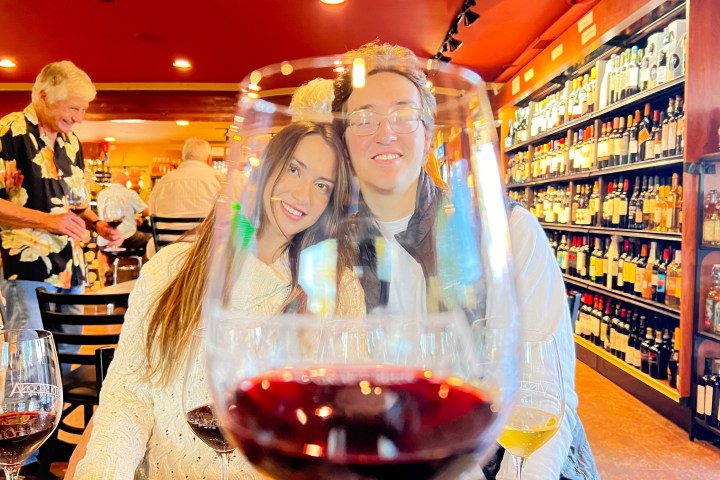 Couple at a wine bar seen through a wine glass, with shelves of bottles in the background.