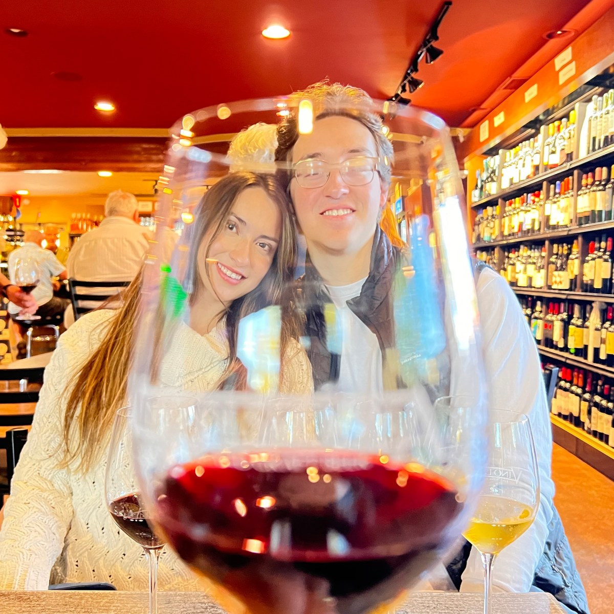 Couple at a wine bar seen through a wine glass, with shelves of bottles in the background.