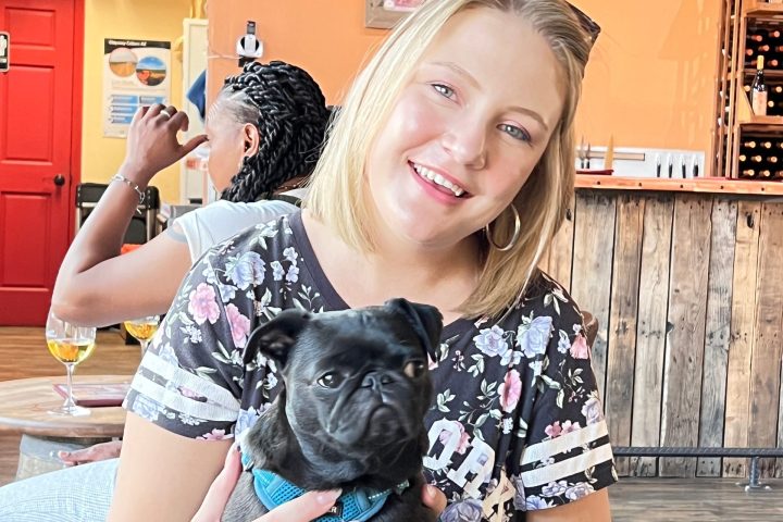 Person holding a black dog in a cafe with a rustic decor and string lights.