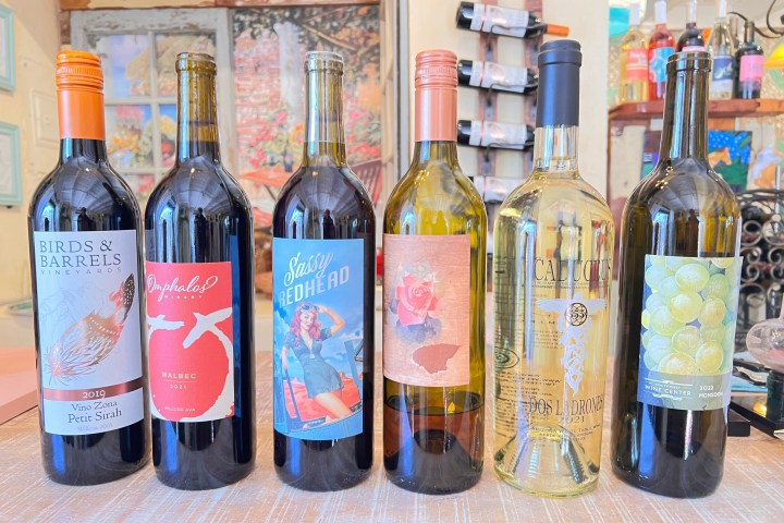 Six wine bottles with colorful labels on a wooden table indoors.