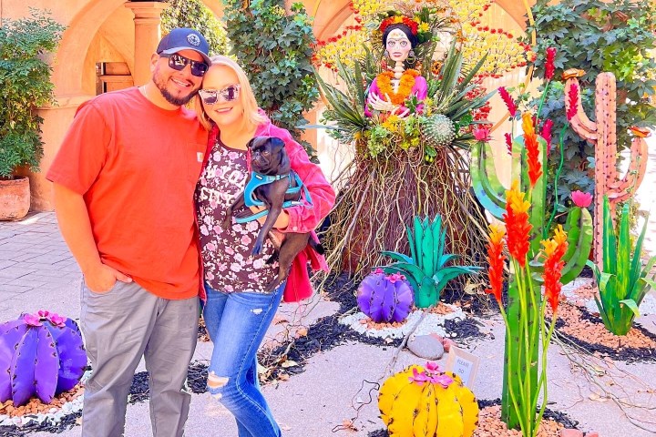 Couple with dog posing near colorful plant decorations and a female-themed sculpture outdoors.