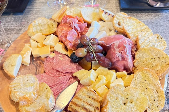 Assorted charcuterie board with meats, cheeses, bread, grapes, and two glasses of white wine.