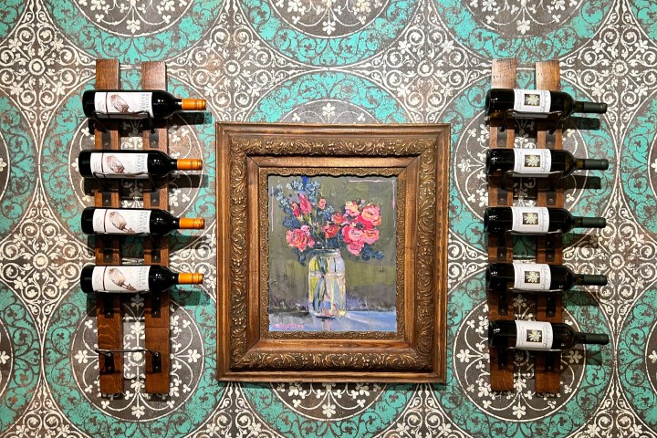 Framed painting of flowers between two wine racks on patterned teal and brown wall.