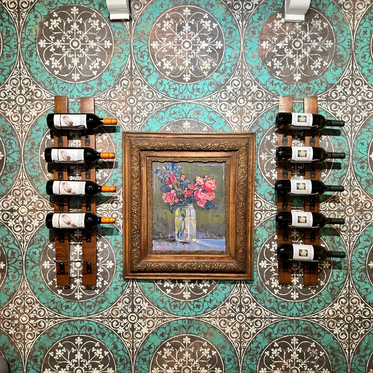 Framed painting of flowers between two wine racks on patterned teal and brown wall.
