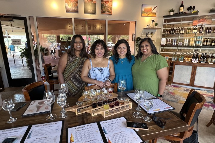 Four women stand smiling behind a table with wine glasses and tasting menus in a cozy restaurant setting.