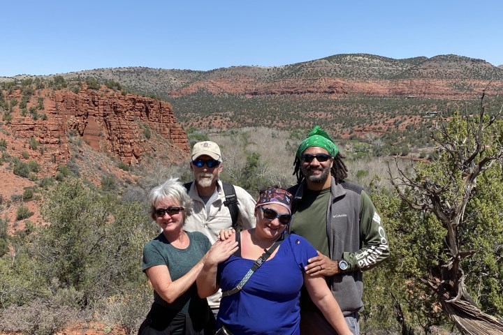 Four people posing in front of red rock cliffs and green shrubs under a clear blue sky.