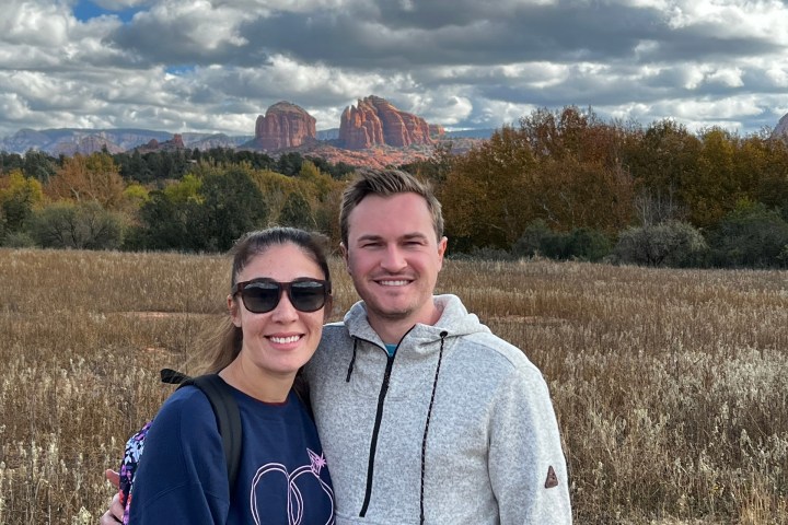 A couple posing in a field with cloudy skies and red rock formations in the background.