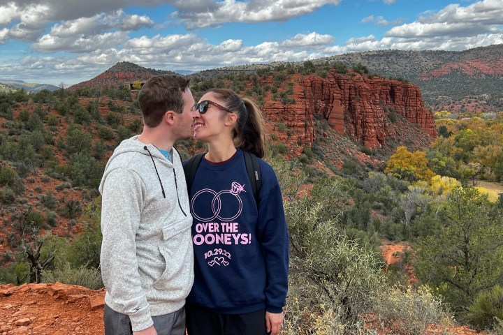 A couple smiles at each other on a red rock trail with cloudy sky and trees in the background.