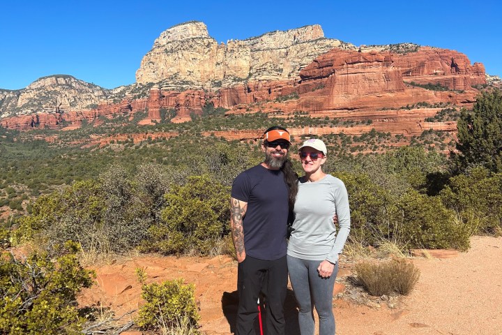 Two people posing in front of a scenic desert landscape with red rock formations under a clear blue sky.