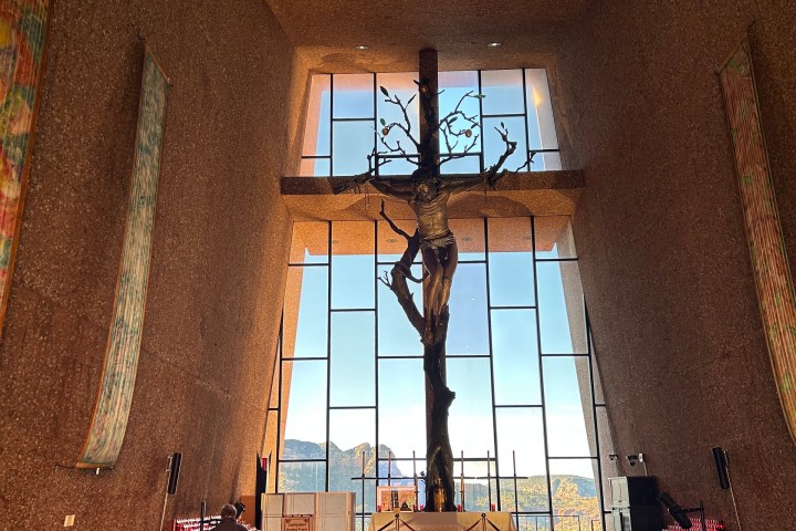 Interior of a chapel with people seated, large crucifix, and bright window in the background.