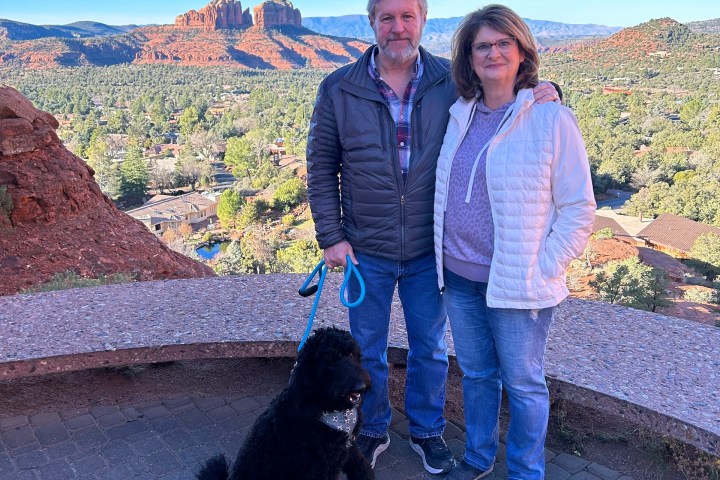 A couple with a black dog standing on a stone path in front of red rock formations.