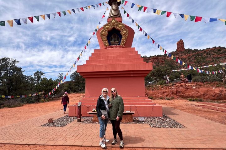 Two people stand in front of a stupa with colorful flags under a cloudy sky.