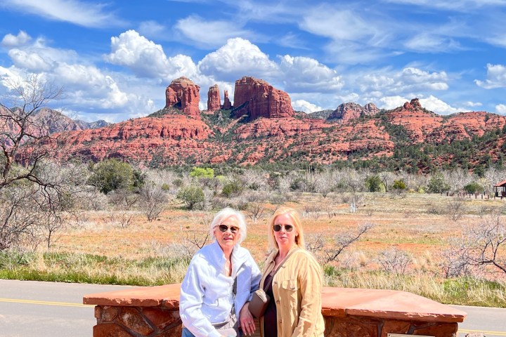 Two people posing in front of red rock formations under a blue sky with clouds in Sedona, Arizona.