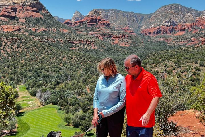 Two people with a dog on a leash stand on a rocky overlook with red rocks and green trees in the background.