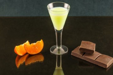 Glass of yellow-green drink, orange slices, and chocolate on a dark reflective surface.