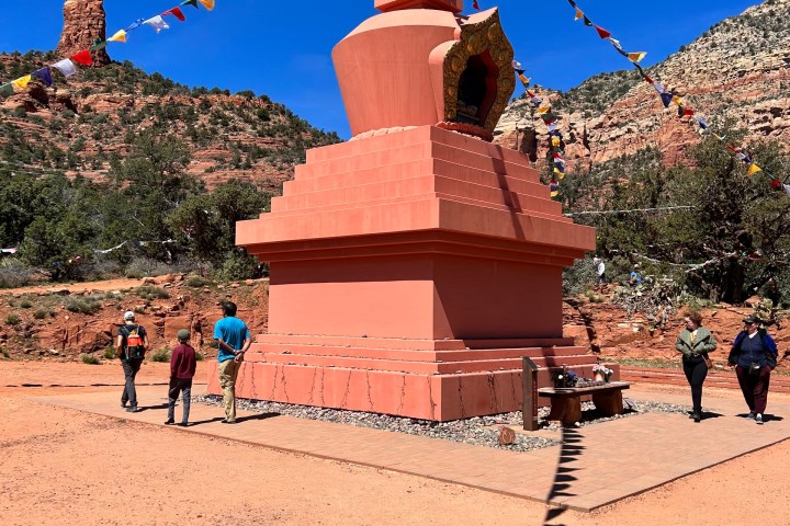 People walking by a Buddhist stupa in a desert landscape with prayer flags and red rock formations in the background.