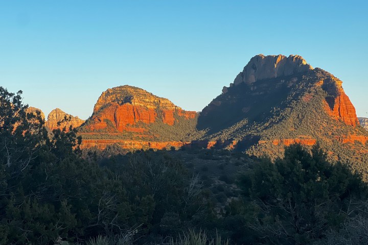 Red rock formations at sunset with clear blue sky and trees in the foreground.