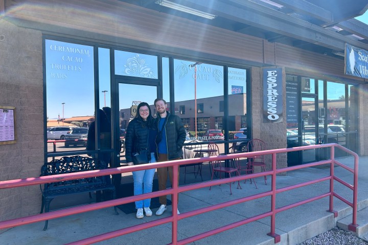 Two people stand smiling outside a cafe with large windows and a red railing on a sunny day.