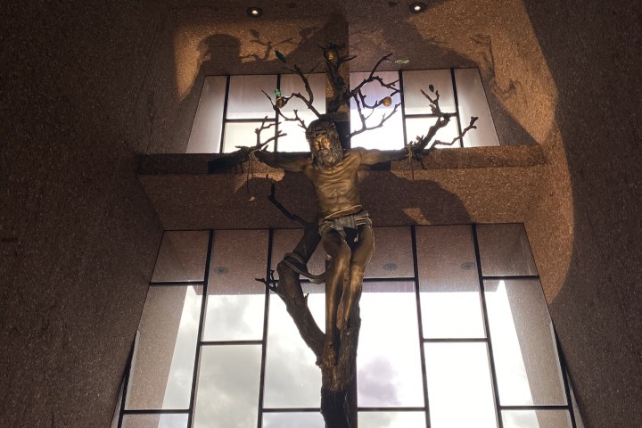 Modern crucifix sculpture against large windows with a cloudy sky and mountains in the background.
