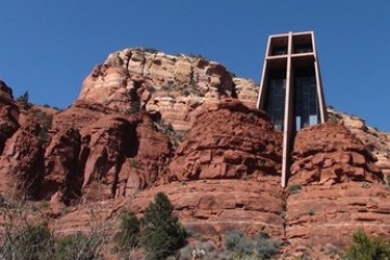 Chapel with a tall cross structure nestled among red rocky cliffs under a clear blue sky.